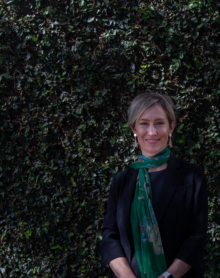A law alumni stands in front of an ivy coloured wall, she is wearing a green scarf and smiling at the camera