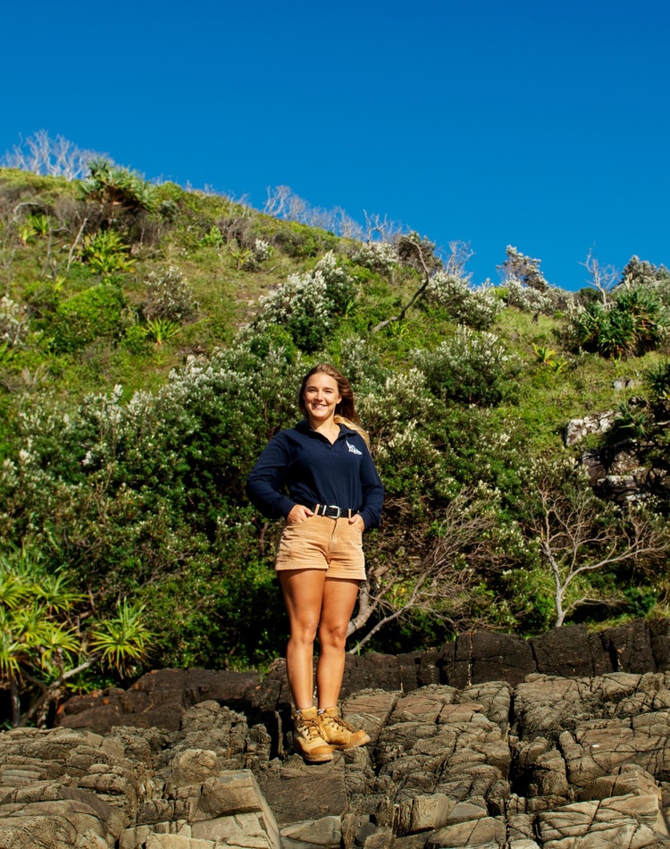 A marine science alumni stands in a rocky coastal landscape, she is wearing a blue long sleeved shirt and brown shorts