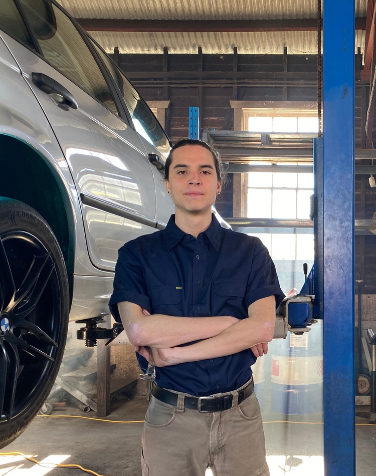 A pathways student is posing in a mechanics workshop , his arms are folded and he has a determined look on his face.