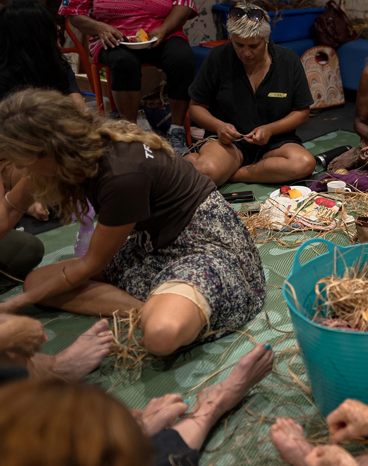 A group of Indigenous and non-Indigenous women sitting on the floor making string from plant fibres.