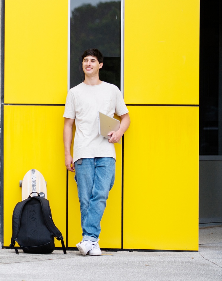 A student leaning on a building with a backpack and notebook.