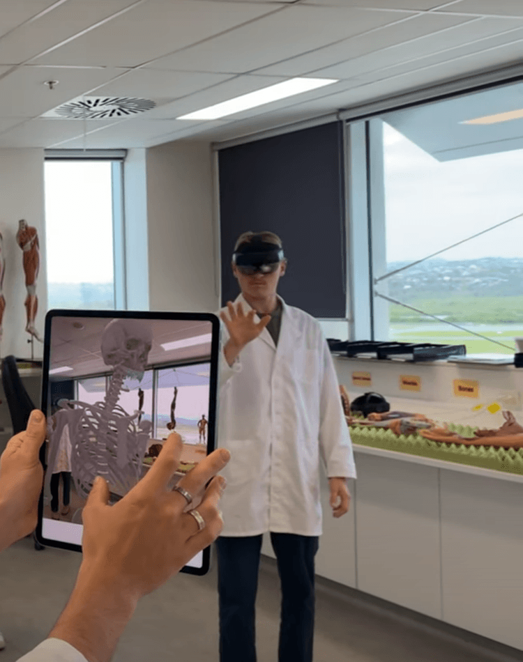 A student holds an ipad, other students wearing goggles in the background