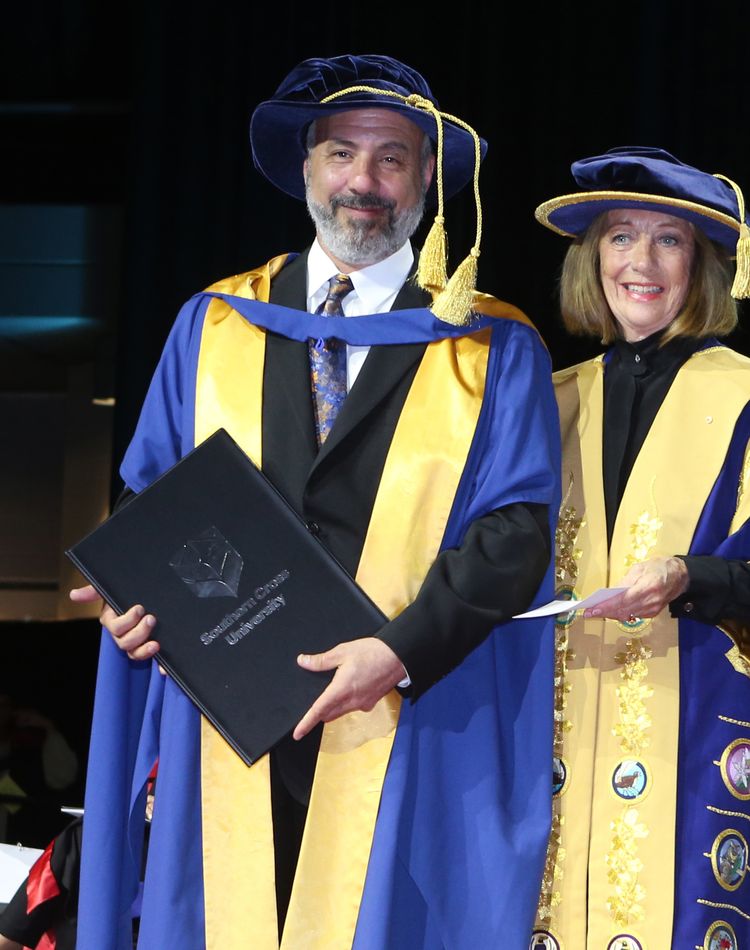 A man and woman in gowns on the dias at graduation ceremony.