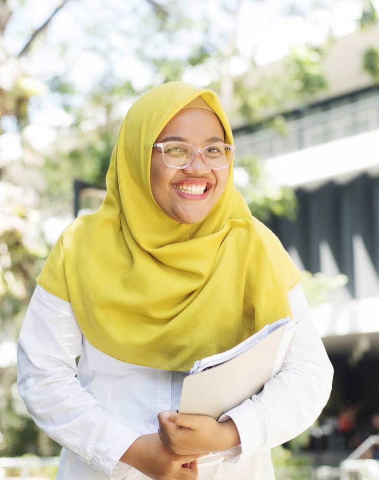 A student with a head scarf and glasses smiling and holding books