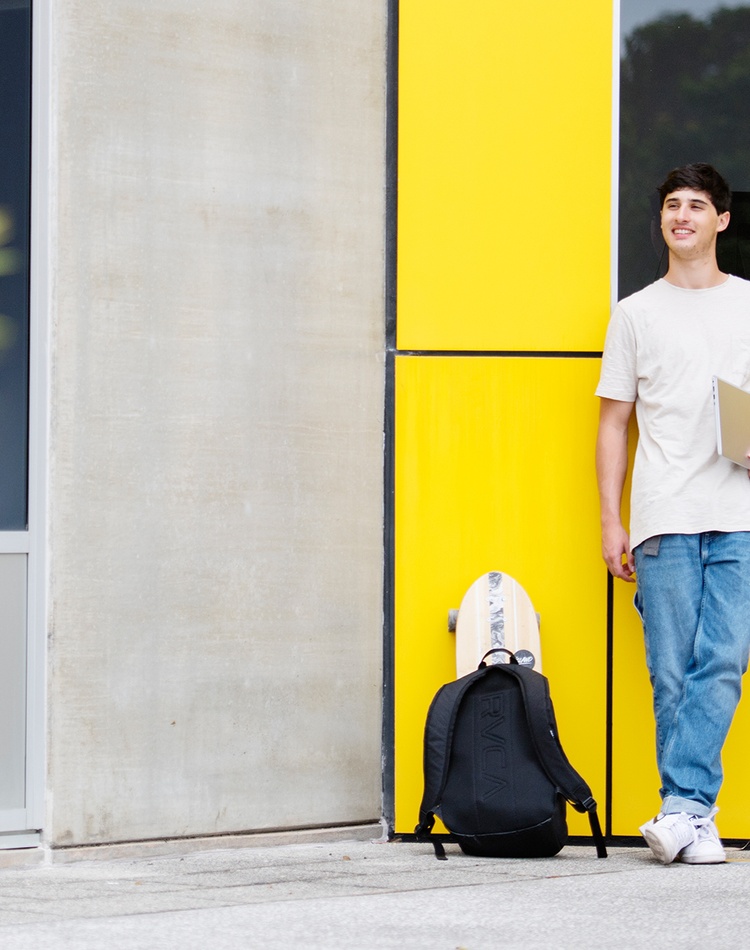 A student leaning on a building with a backpack and notebook.