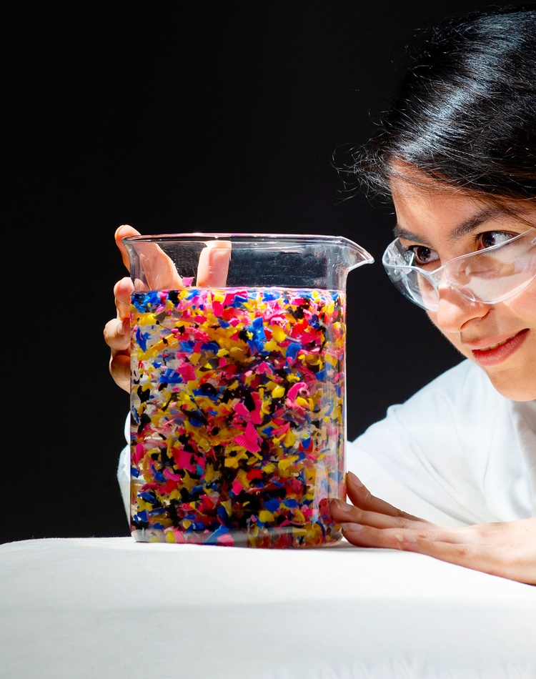 Dr Priya Borpatra Gohain BSc, MSc, PhD (SCU)  in a white lab coat looking into a large beaker or water filled with brightly colour coloured plastic particle's at Southern Cross University Lismore Campus