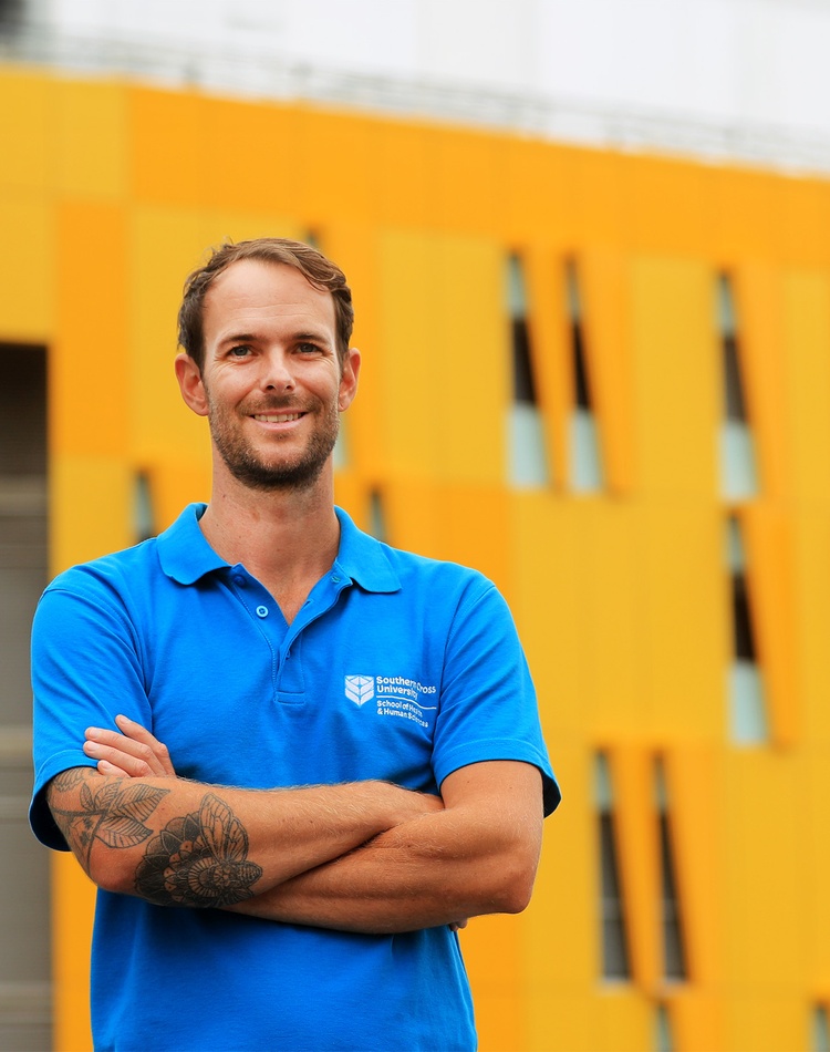 Beau Halligan graduated from the Master of Osteopathy in 2021, he is standing in front of the southern cross university gold coast campus wearing a blue shirt with his arms crossed