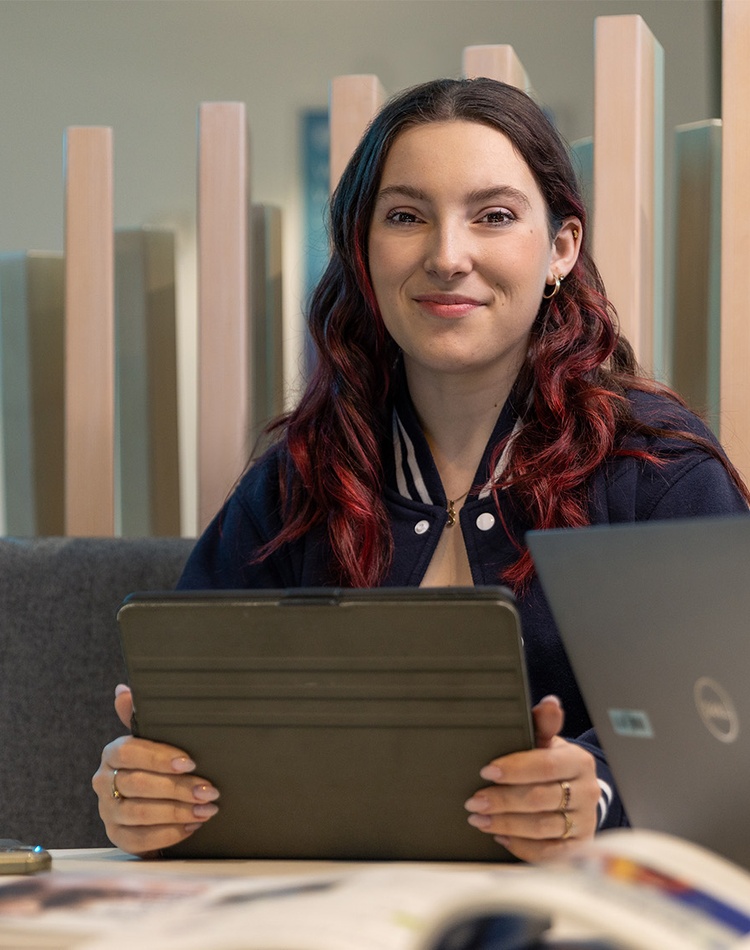 Student with iPad in library and laptops on her left and right