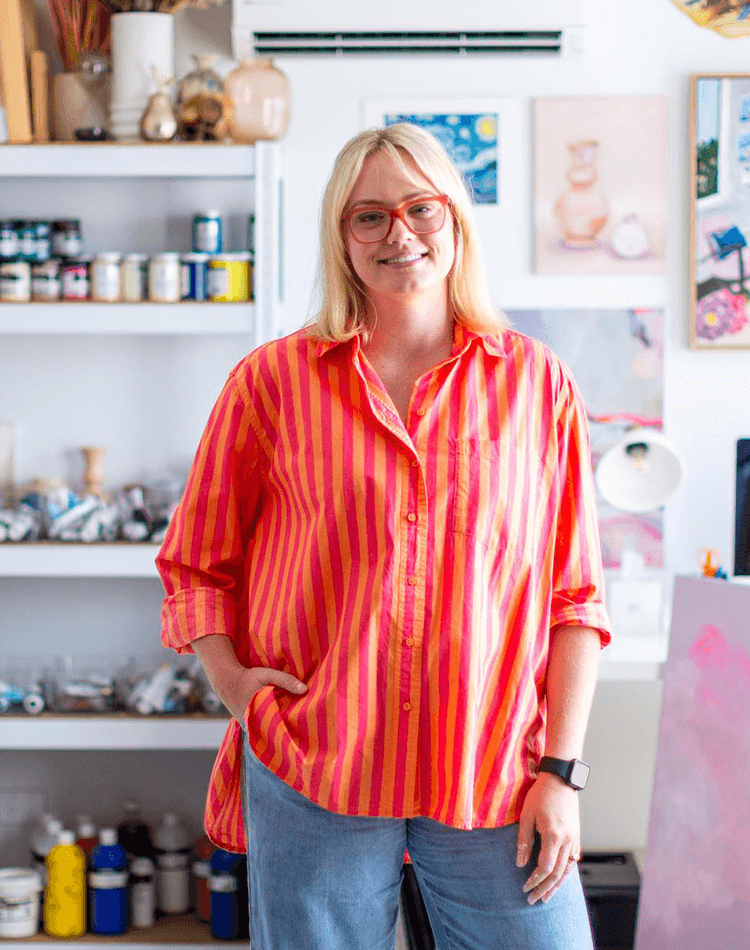 Woman in bright striped shirt standing in art studio with supplies and canvases in background