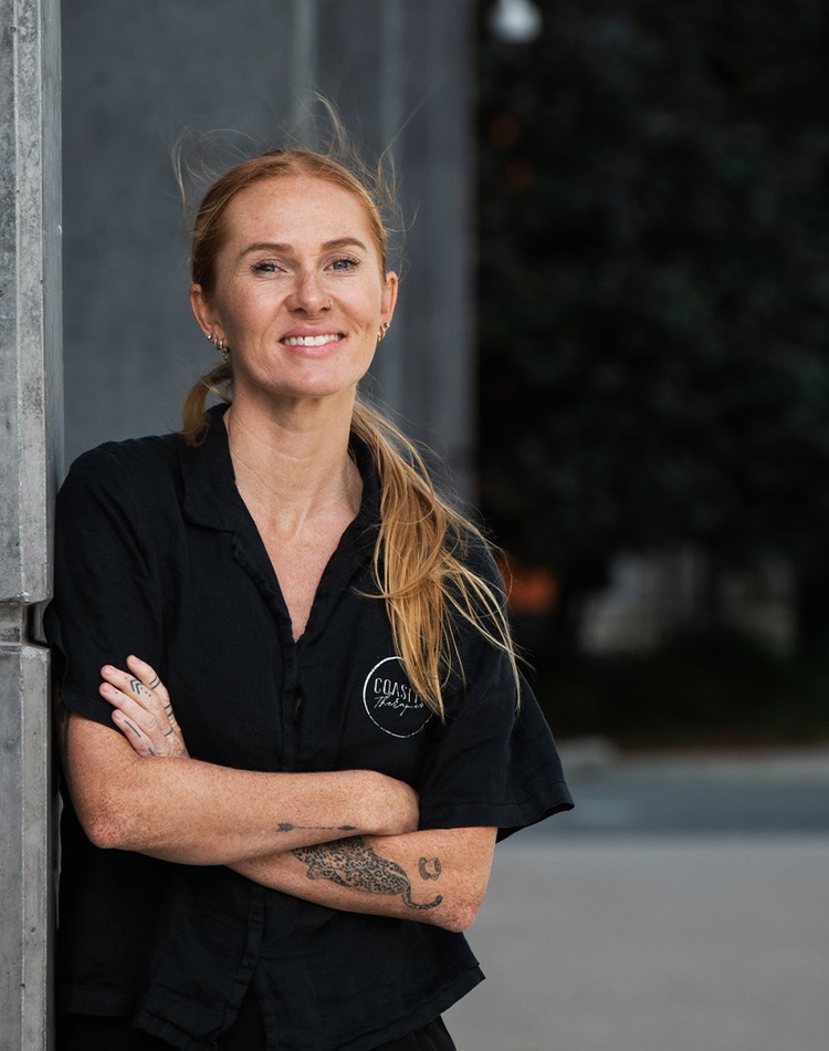Woman in navy scrubs smiling with arms crossed, leaning against cement wall