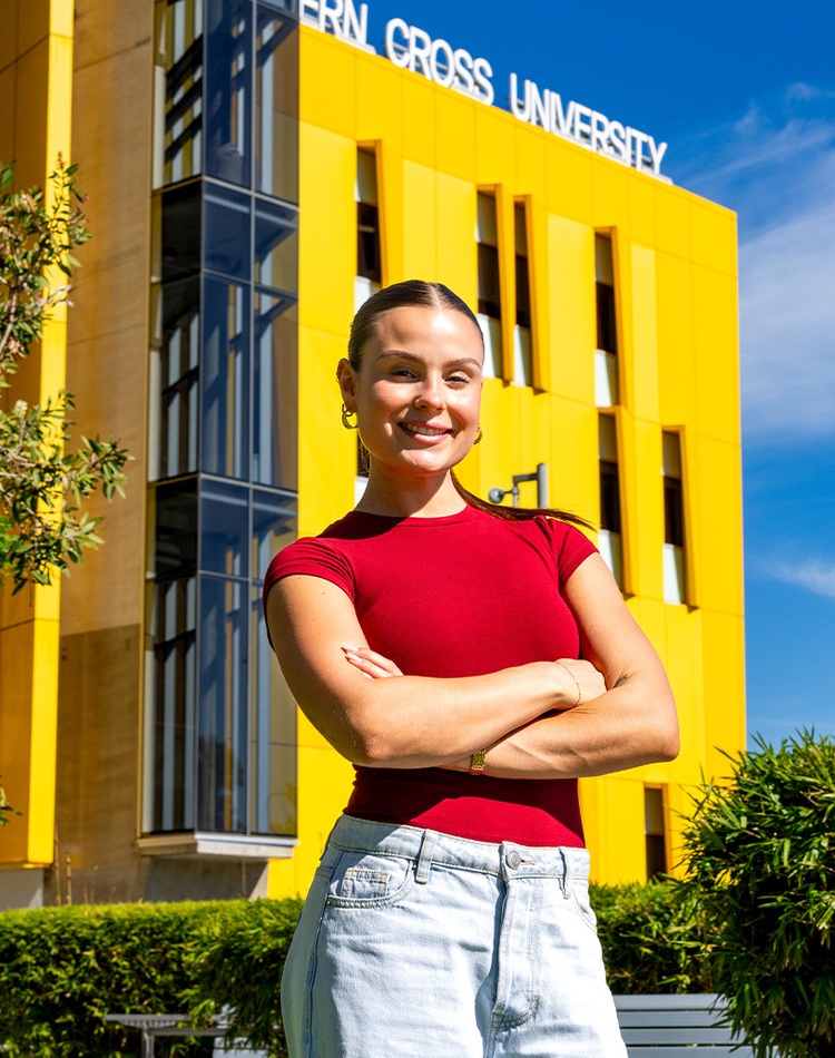Woman in red shirt smiling with arms crossed in front of yellow building with Southern Cross University written on the roof