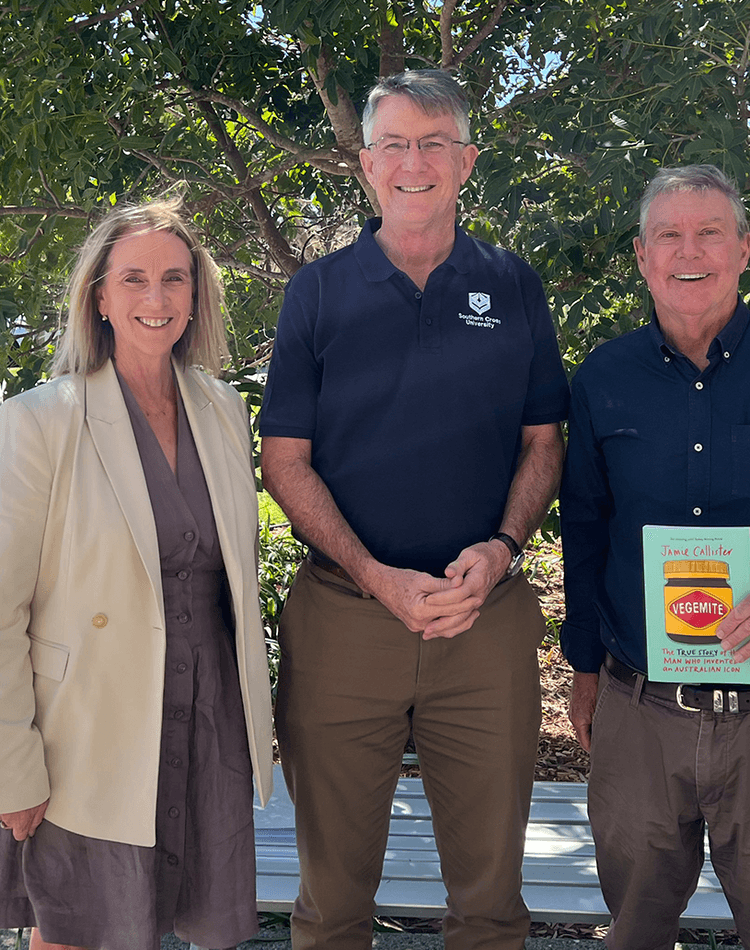 Three people posing in front of greenery, one holding a book with a jar of vegemite on the cover