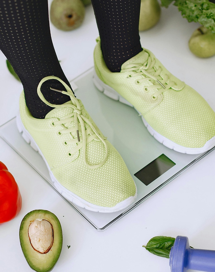 Person standing on weight scales surrounded by tape measure and fresh vegetables