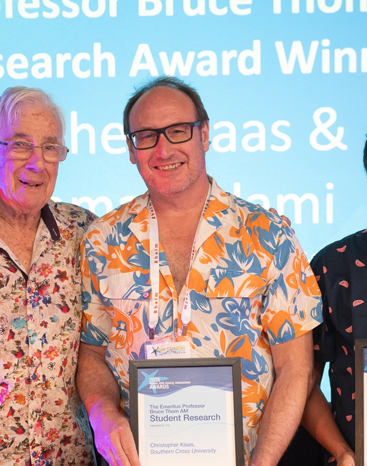 3 men on a stage in front of a screen, 2 of them holding awards