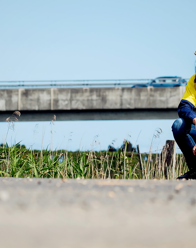 An engineering alumni squats down in front of a concrete bridge, she is wearing a high visibility yellow safety hat and high visibility shirt