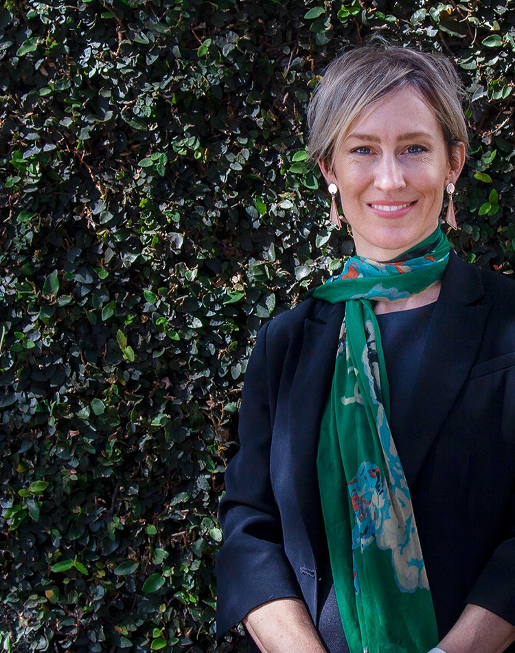 A law alumni stands in front of an ivy coloured wall, she is wearing a green scarf and smiling at the camera