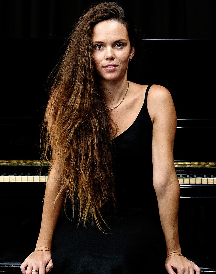 A music student is sitting in front of a piano on a stool she is smiling at the camera and has long hair