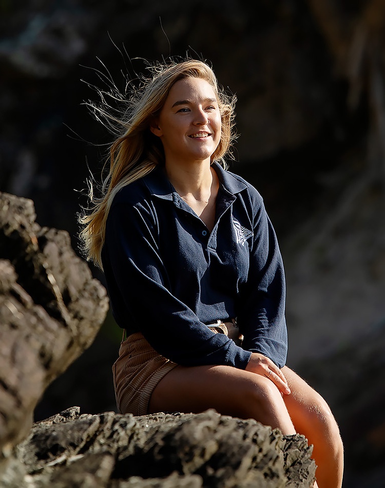 A marine science alumni sits in a rocky coastal landscape, she is wearing a blue long sleeved shirt and brown shorts and is smiling off to the side of the camera