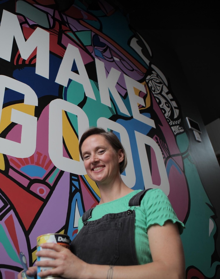 An arts alumni standing in front of a mural she is paining, she is holding a can of paint and smiling at the camera