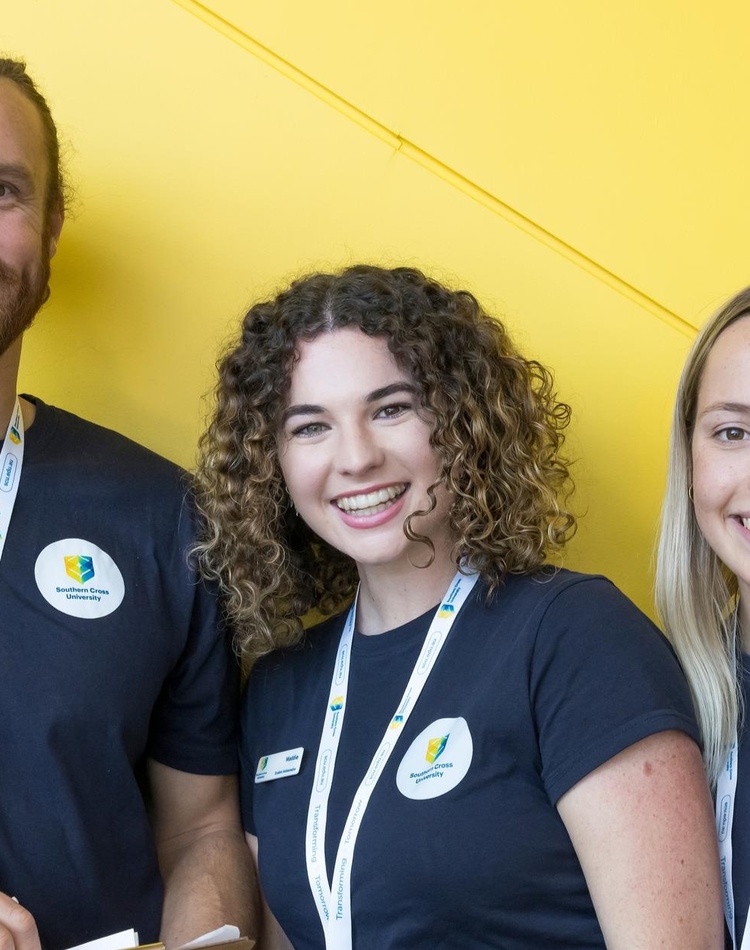 Three student volunteers smile at the camera in front of a yellow wall.