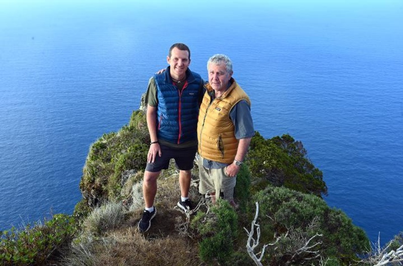 Two men standing on a mountain top with ocean in the background