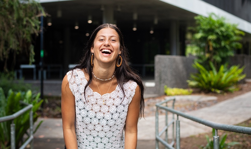 Pearl Andrews, Bachelor of  Indigenous Knowledge student stands laughing outside the Lismore Library at Southern Cross University surrounded by trees and native plants