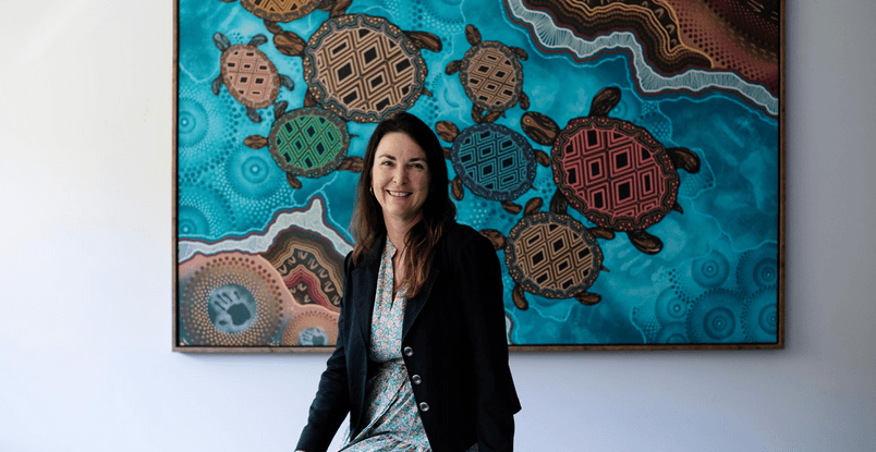 Doctor of Indigenous Philosophies graduate Kylie Day sitting in front of painting