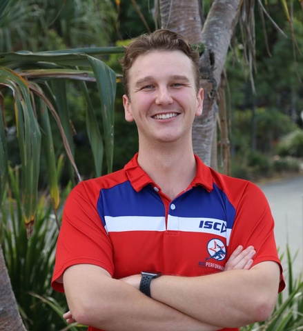 Headshot of person in red shirt with arms crossed and greenery in background