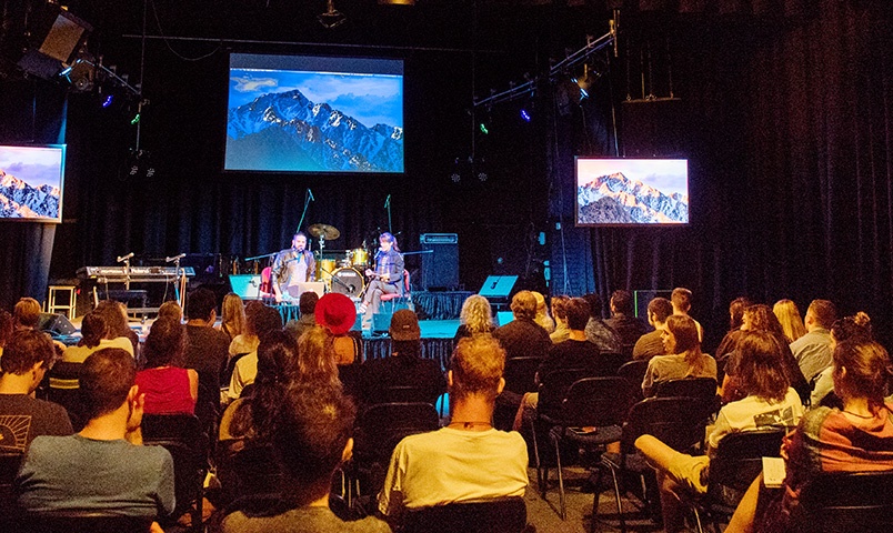 Audience sits watching a musical performance with two performers on stage and three screens showing mountain ranges behind them