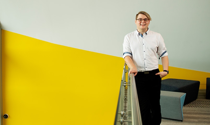 Owen Lednor, Director of Care at The Shoreline Luxury Retirement Living Centre and SCU graduate stands in their offices in front of a bright yellow wall with comfortable seating behind him