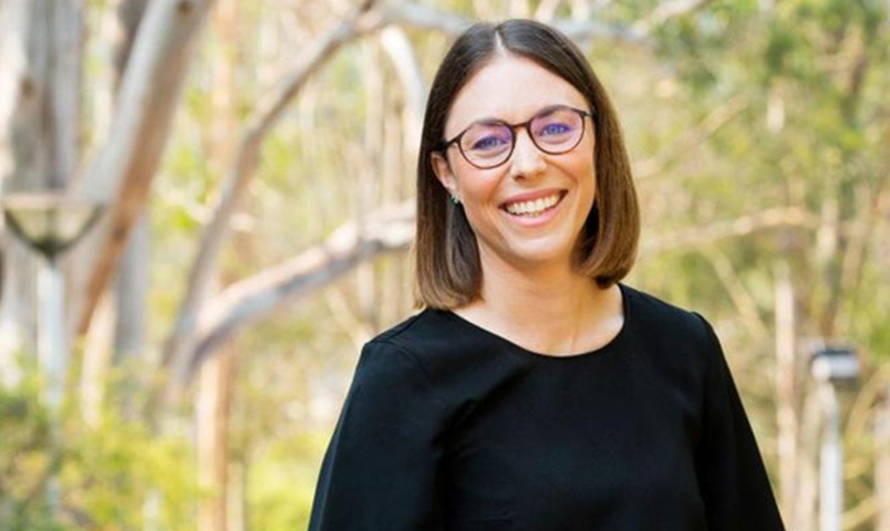 Natasha Cook, SCU graduate smiles outside in front of gumtrees wearing a black dress and black glasses