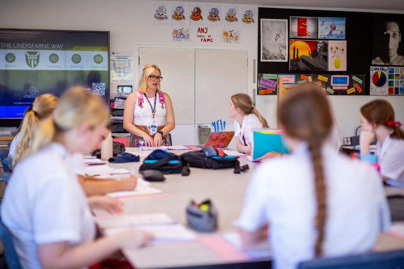 A teacher stands at the front of a classroom, the students listen closely to what she is saying.