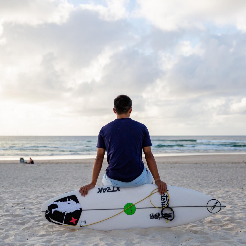 A student sitting on the beach, leading against a surfboard