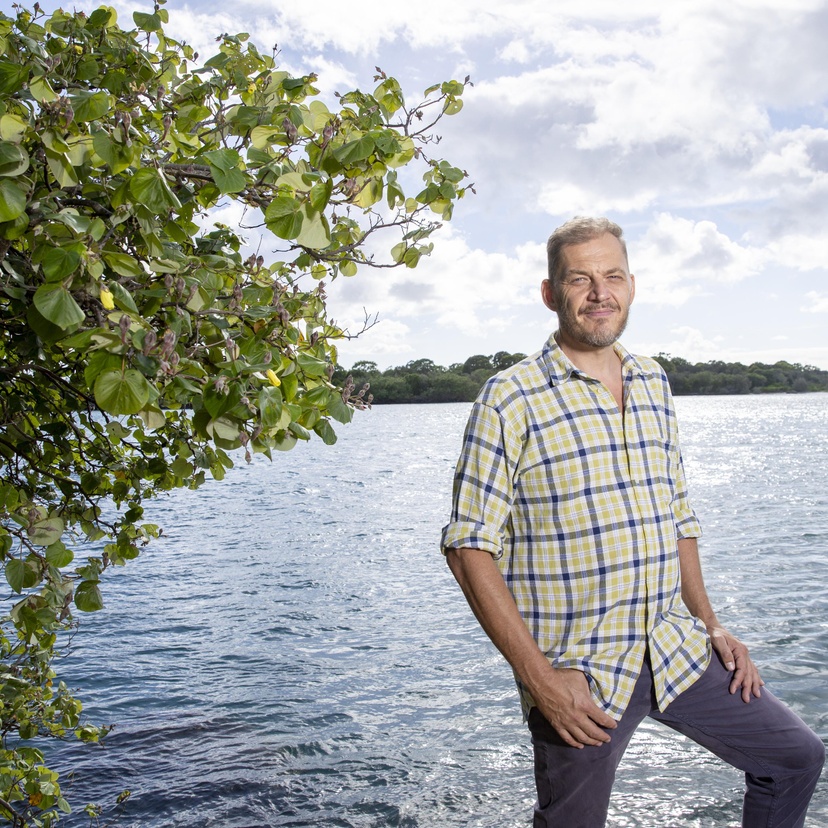 a social work post graduate stands next to some mangroves and a bay, he is looking into the camera