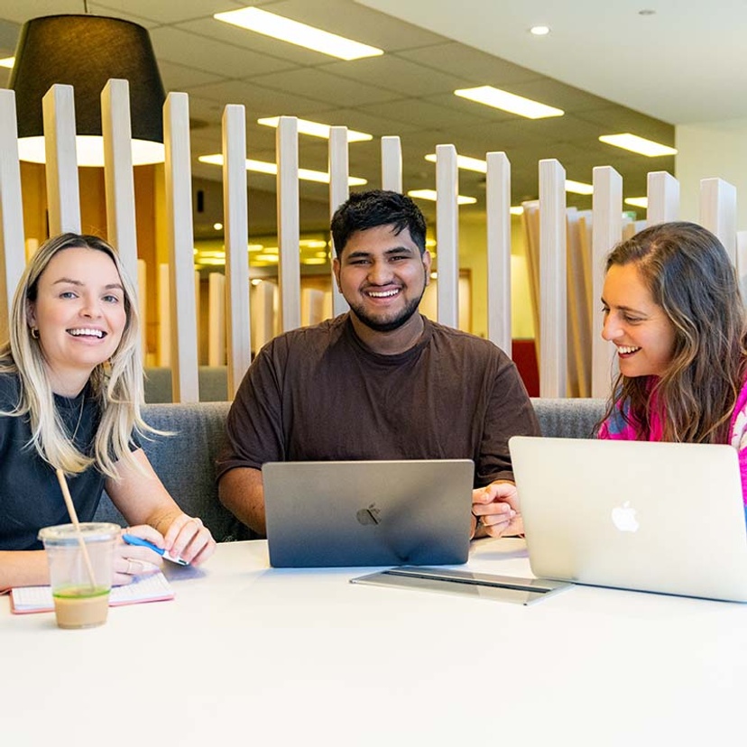 Three students smiling and sitting at table with laptops in the Gold Coast campus library