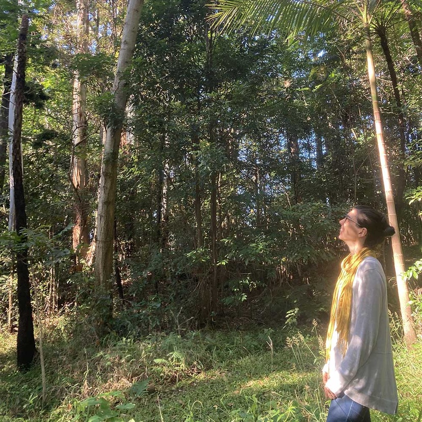 person white top and blue jeans, standing looking up at trees and smiling