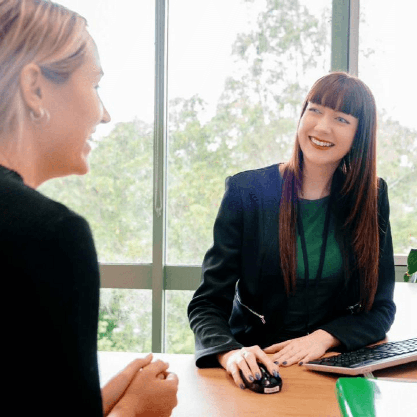 Two ladies sitting across the desk from one another smiling
