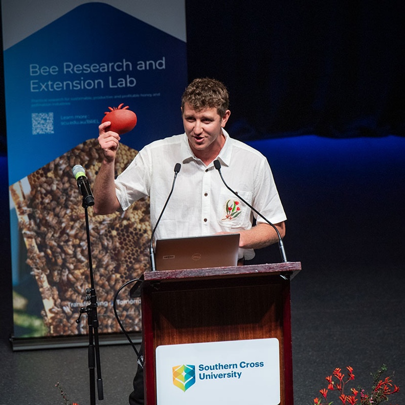 Dr Cooper Schouten standing at podium holding a piece of fruit (pomegranate)