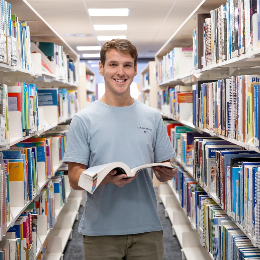 person smiling looking at camera standing in front of shelves of books, holding an open book