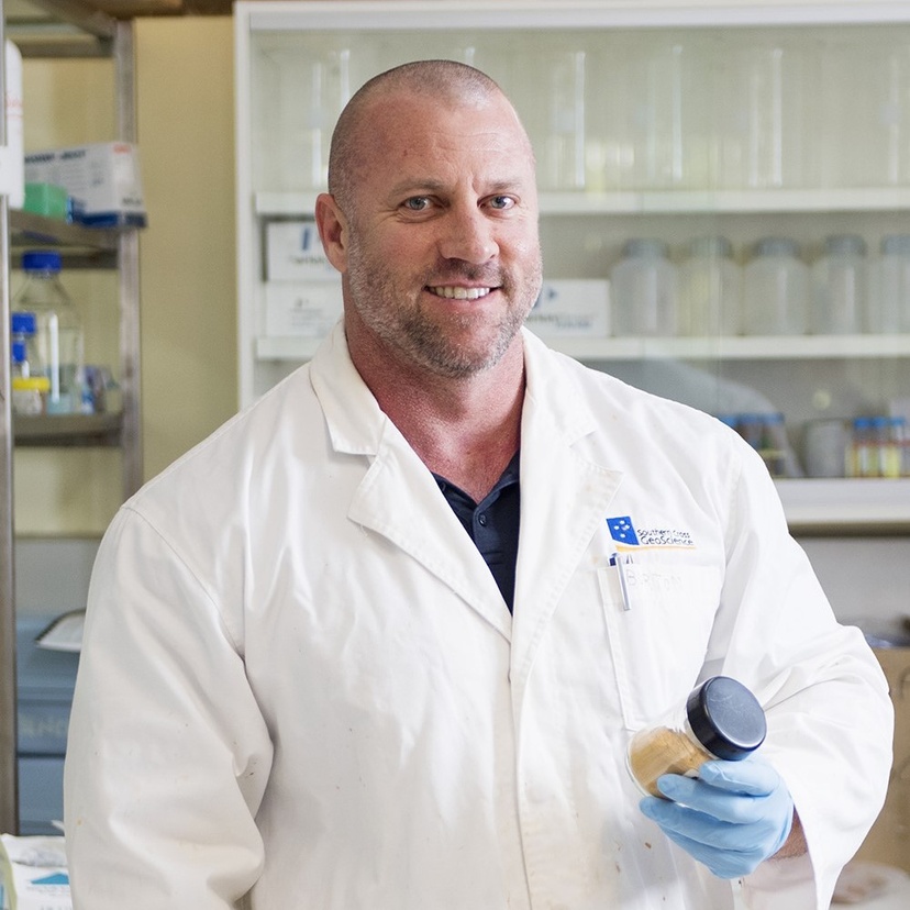 Man smiling at Camera wearing white lab coat and blue gloves holding a jar