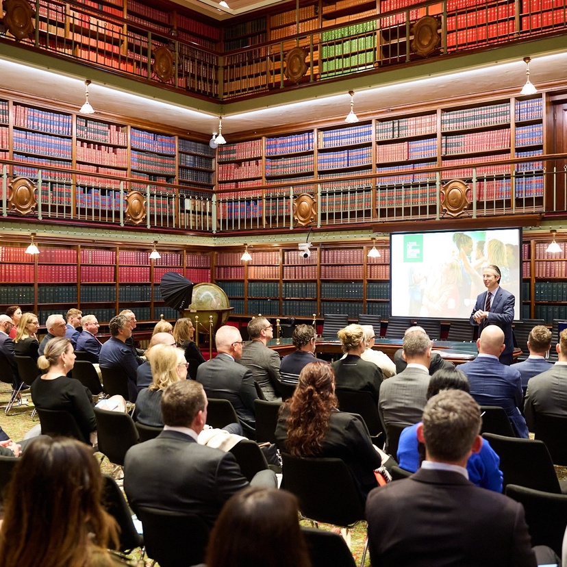 Room with shelves full of books, man standing at front of room in front of many seated people