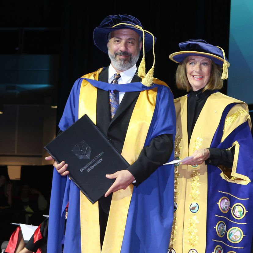 A man and woman in gowns on the dias at graduation ceremony.