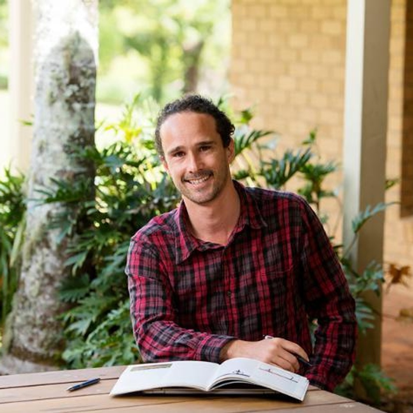 A man with a beard in a red check shirt sitting at a table with a textbook