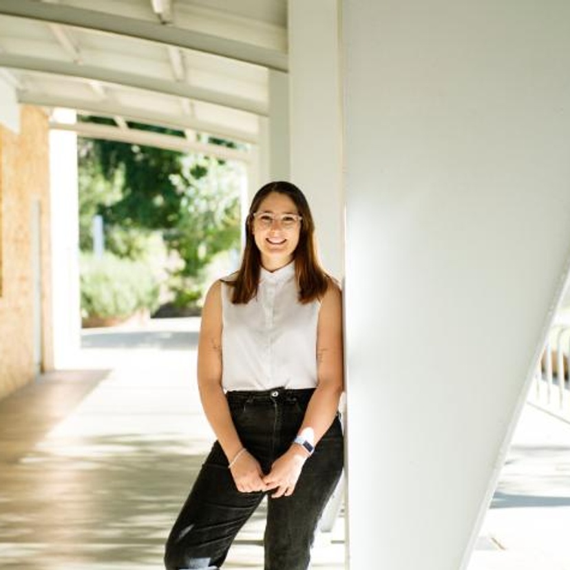 Woman in white shirt and black jeans outside leaning against a building structure