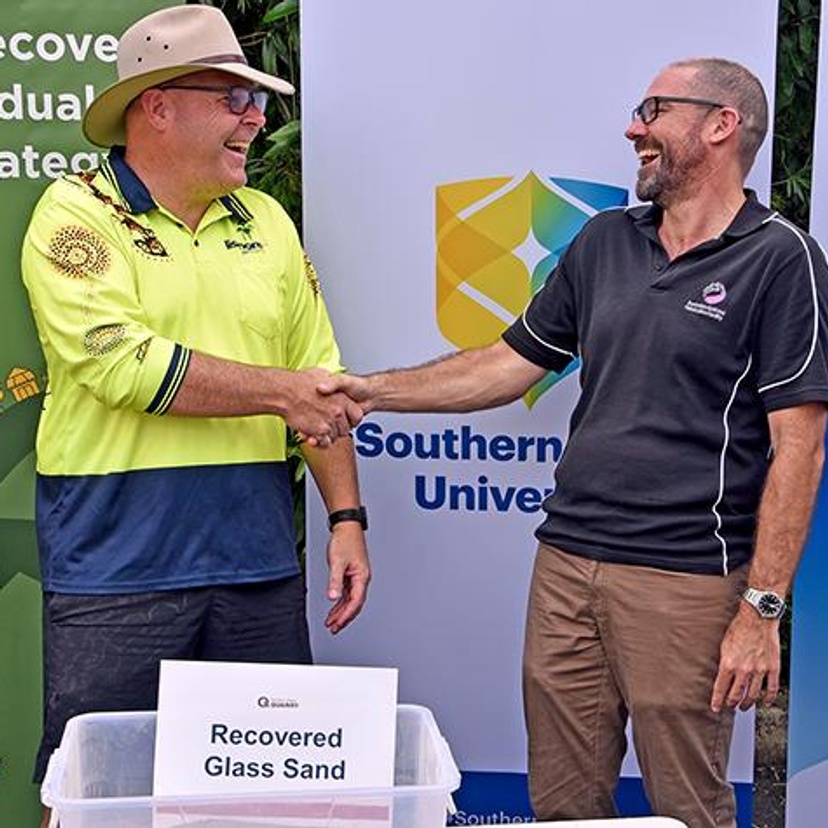 Two men shaking hands in front of a container of recycled glass