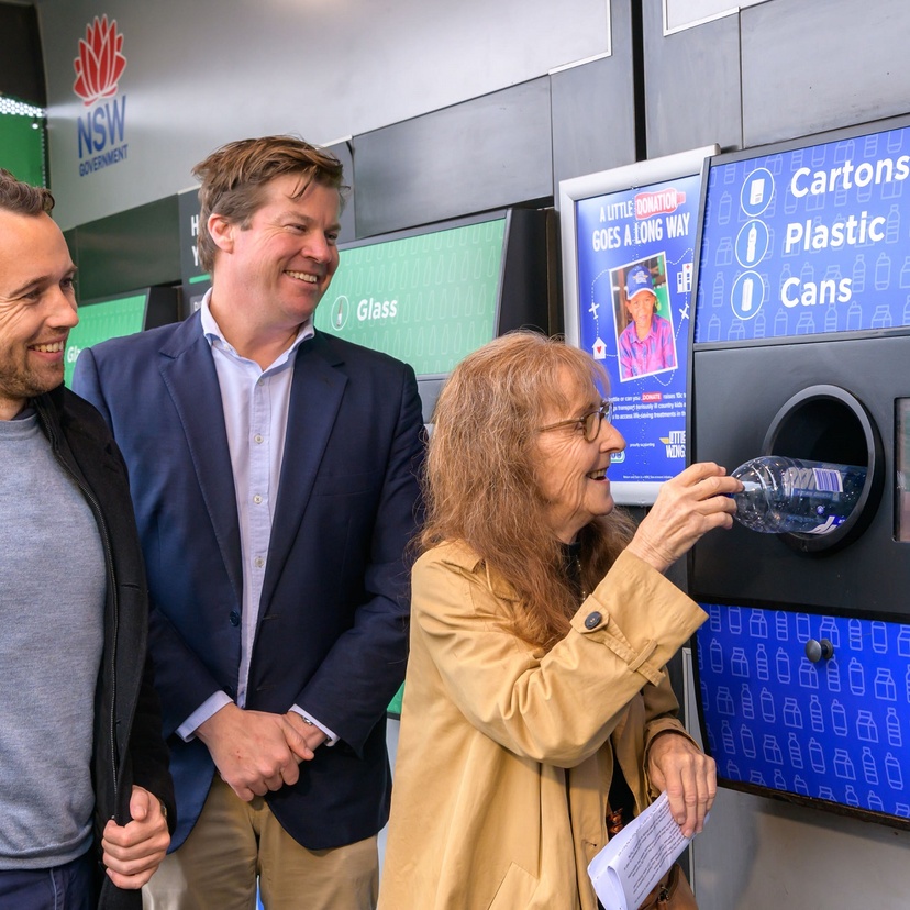 Two men watch as a woman feeds a plastics bottle into a recycling station
