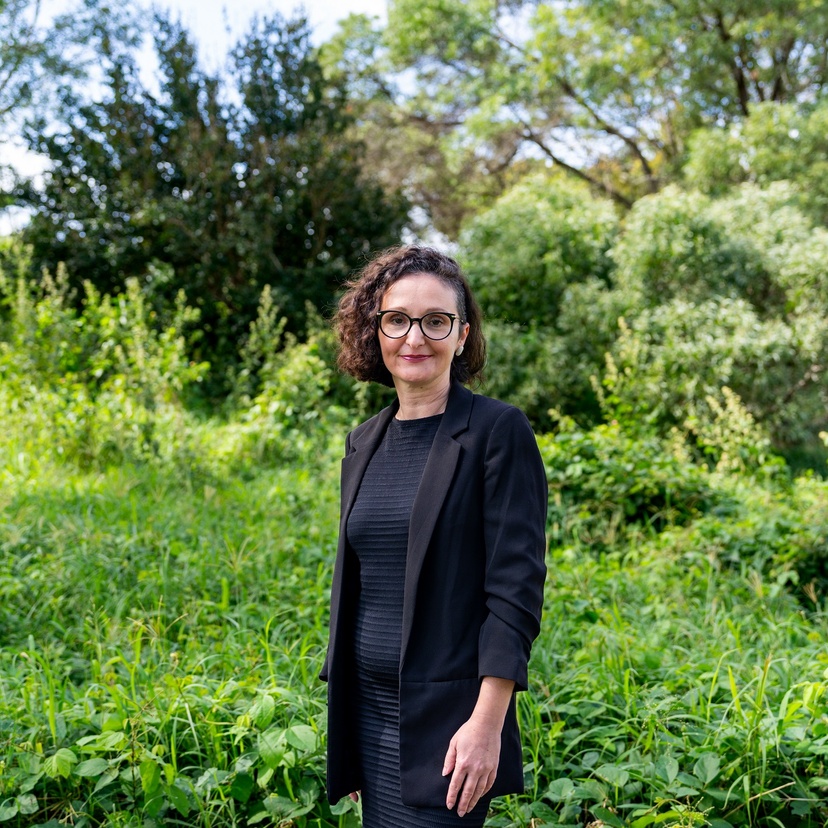 A woman faces the camera, greenery in the background