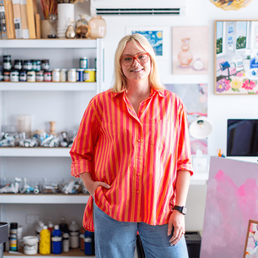 Woman in bright striped shirt standing in art studio with supplies and canvases in background