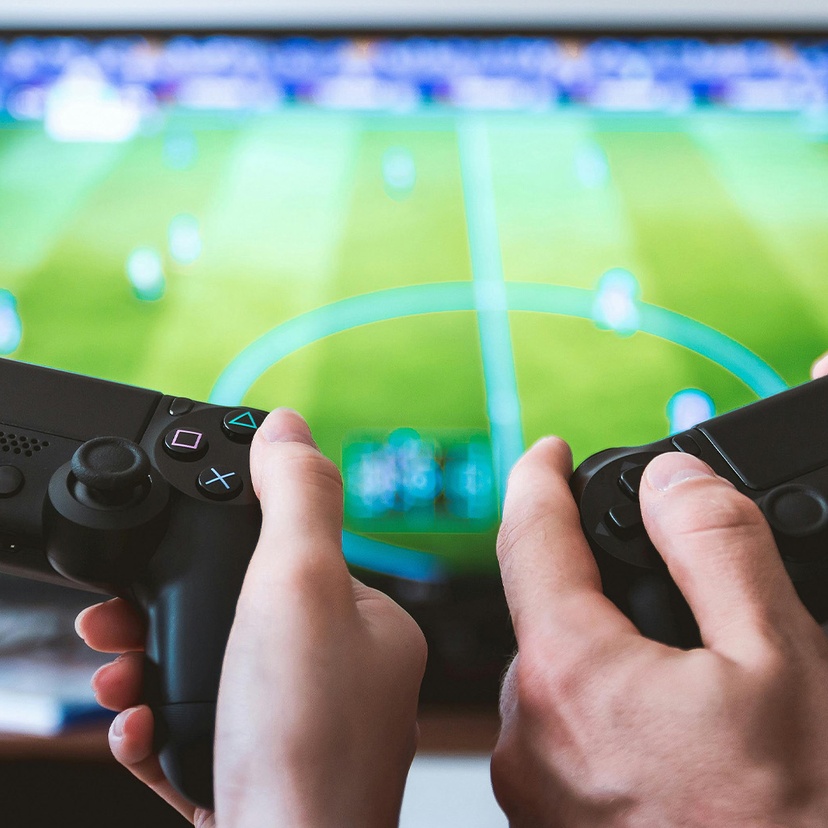 Twos sets of hands holding PlayStation 4 controllers with tv with sports game in background