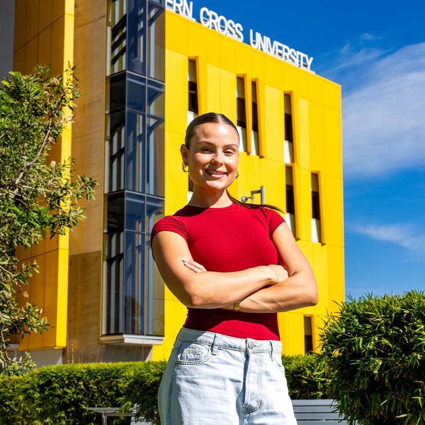 Woman in red shirt smiling with arms crossed in front of yellow building with Southern Cross University written on the roof